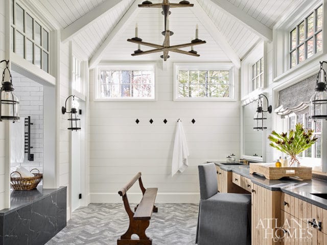 White shiplap walls and vaulted ceiling with wood beams in a room with herringbone tile floors, a wooden bench, and a vanity with wood cabinetry and a stone countertop.