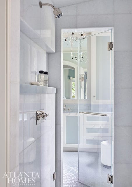 White marble bathroom with glass shower enclosure, vanity with frosted glass cabinets, and a crystal chandelier