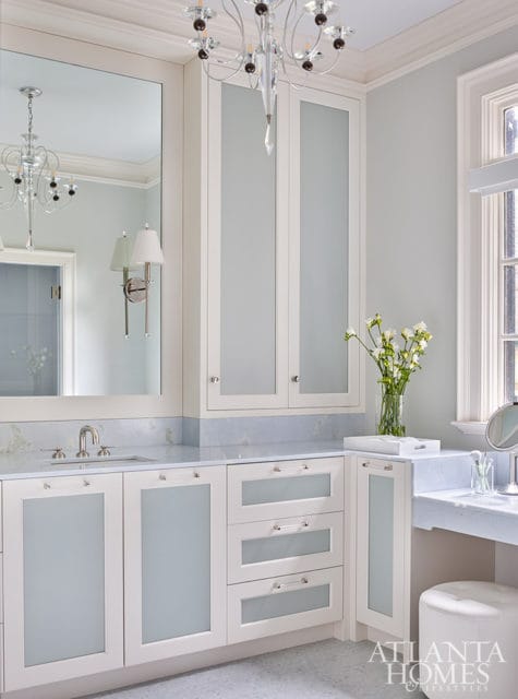 Light blue and white bathroom with white cabinetry, frosted glass doors, marble countertops, and a crystal chandelier.