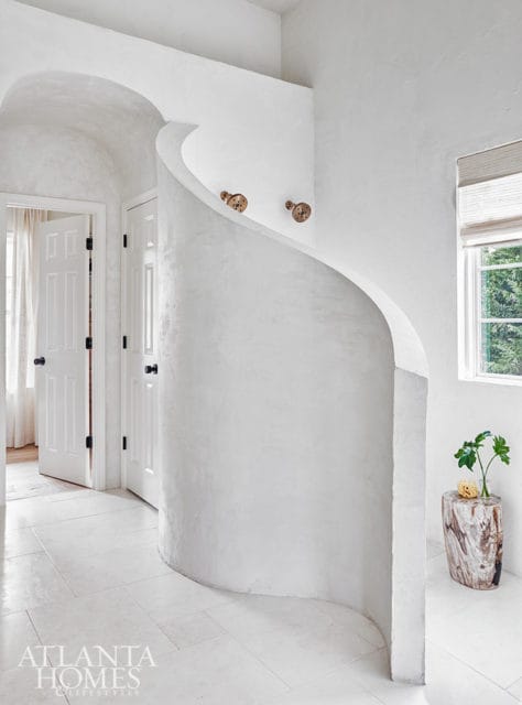 White hallway with curved plaster wall, two doors, and a window overlooking greenery. A log-shaped side table holds a vase with a plant.