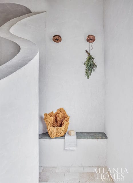 Spa-like bathroom with curved white plaster walls, a stone bench with a large natural sponge and folded towel, and a bundle of dried herbs hanging from the ceiling.