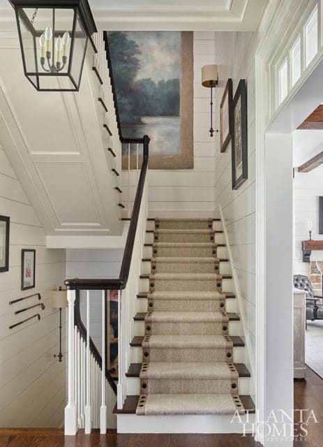 Staircase with shiplap walls, a landscape painting, and a patterned runner with dark trim and nailhead accents. A black lantern hangs overhead.