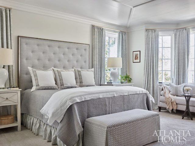 Bedroom with a tufted gray headboard, gray bedding, white pillows with decorative trim, and patterned curtains. A small sofa and a patterned ottoman are visible.