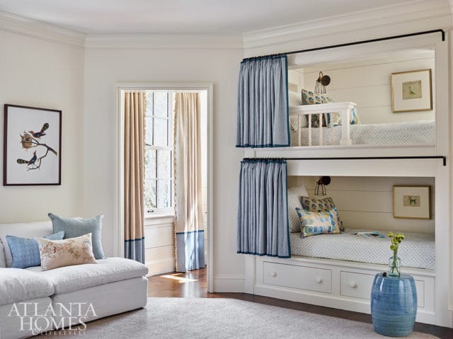 Built-in bunk beds with blue curtains, white shiplap walls, and framed artwork. A white sofa with patterned pillows sits in the foreground next to a window with beige and blue curtains.