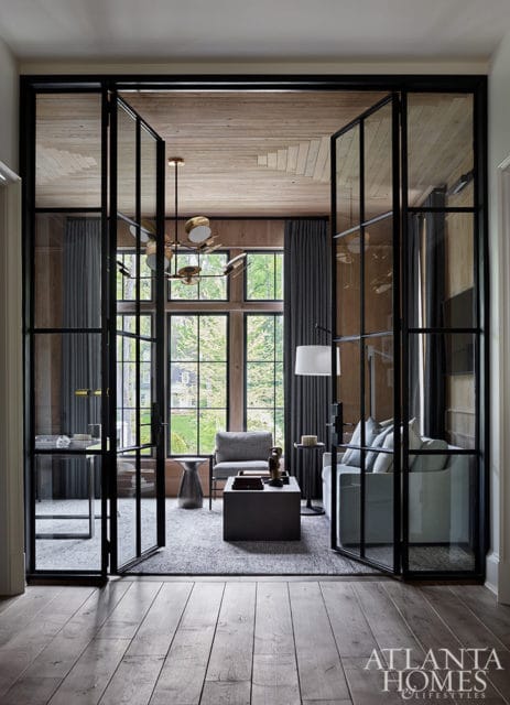 Modern living room with glass-paneled doors, light wood ceiling, gray upholstered furniture, and a large window overlooking greenery.