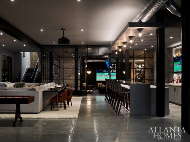 Modern basement entertainment area with a long sectional sofa, bar seating, exposed brick walls, and a large screen displaying a baseball game.