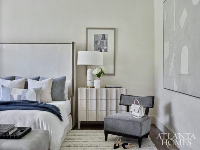 Bedroom with a white upholstered headboard, gray and white patterned pillows, a navy throw blanket, a fluted white nightstand with a white lamp and vase, and a gray accent chair with a white handbag.