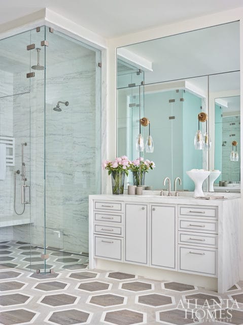 White bathroom vanity with marble countertop, double sinks, and mirrored cabinets. Features a glass-enclosed shower with marble tile walls and a hexagonal floor pattern.