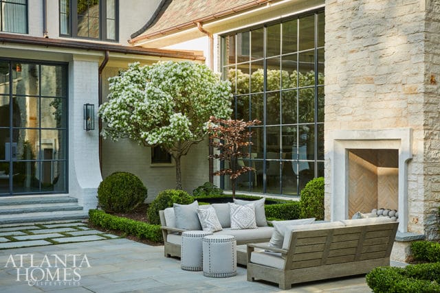 Stone patio with light gray outdoor sofa and chairs, two studded ottomans, and a blooming tree. Large grid windows reflect the outdoors.