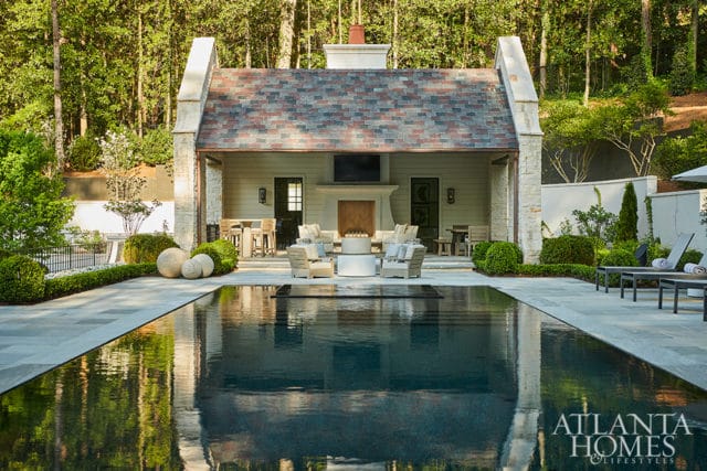 Modern outdoor living space with a dark-bottomed swimming pool, stone patio, and covered pavilion featuring a fireplace and seating area, surrounded by lush landscaping and trees.