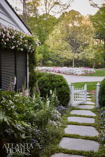 Stone pathway leading to a white picket gate, flanked by lush gardens with pink and white flowers, and a house with climbing pink roses.