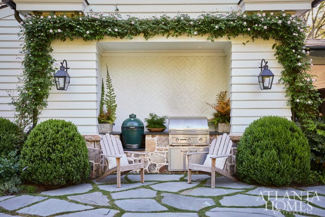 Outdoor kitchen with built-in grill and green egg smoker, flanked by Adirondack chairs and lush greenery, under a vine-covered pergola.