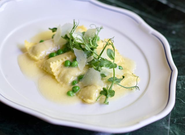 Ravioli with peas and shaved parmesan cheese, garnished with microgreens, served in a light butter sauce on a white plate with a blue rim.