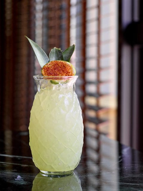 Pineapple-shaped glass filled with a pale yellow cocktail, garnished with a lime slice dusted with chili powder and pineapple leaves.