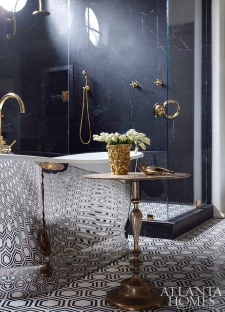 Master bathroom with black marble walls, freestanding tub with patterned skirt, gold fixtures, glass shower enclosure, and geometric floor tiles.