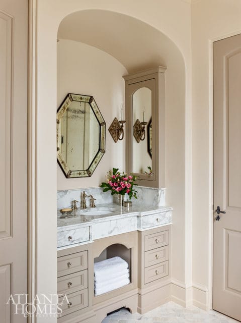 Bathroom vanity with marble countertop, brass fixtures, and taupe cabinetry. An octagonal mirror hangs above the sink, with a tall mirrored cabinet to the right.
