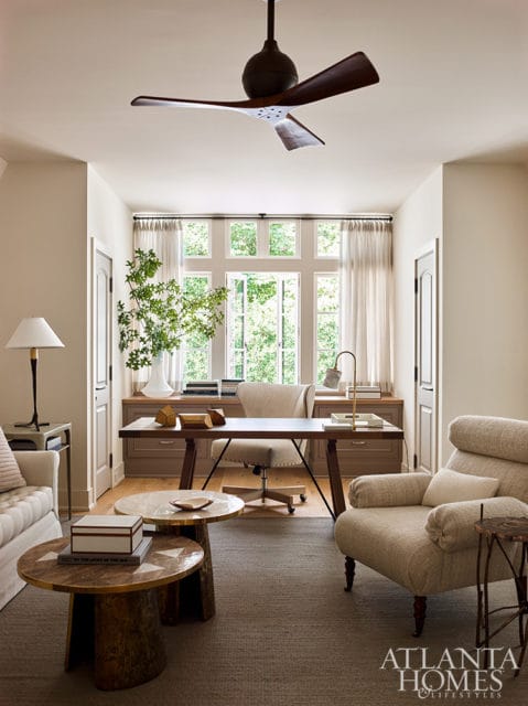 Home office with a large desk and chair facing a wall of windows, flanked by built-in cabinetry. A sofa and armchair are visible in the foreground.