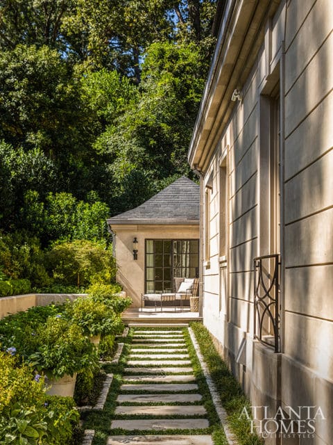 Stone pathway leading to a covered patio with a glass door and seating, flanked by lush greenery and a light-colored stone building.