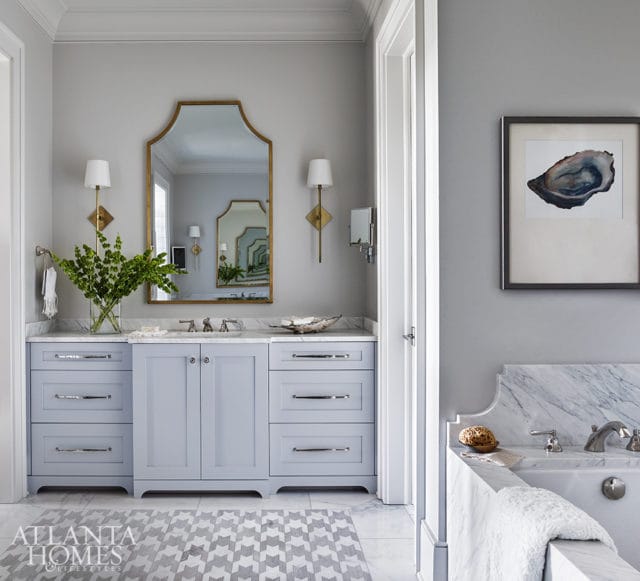 Light blue bathroom vanity with marble countertop, brass sconces, and a gold-framed mirror above. A framed oyster print hangs on the gray wall.