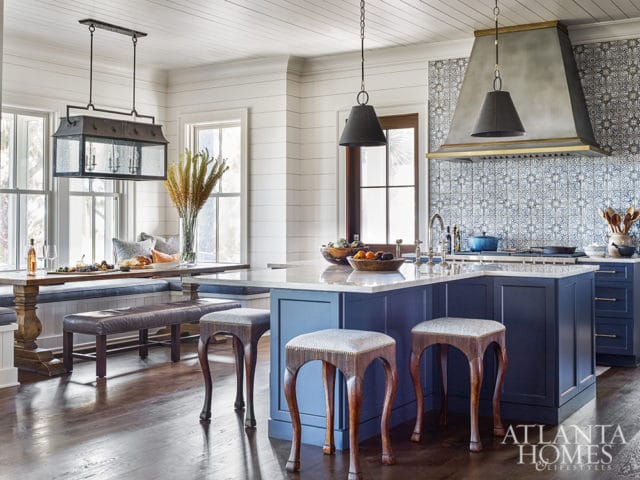 Blue kitchen island with marble countertop and dark wood stools, adjacent to a dining area with a wooden table, leather benches, and a large window.