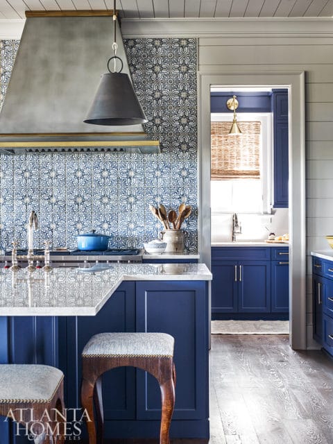 Blue kitchen with patterned tile backsplash, marble island, and dark metal pendant light. A doorway reveals a second blue kitchen area.