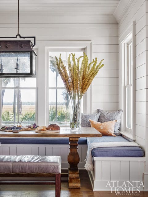 Dining nook with shiplap walls, built-in banquette seating with blue cushions, a wooden table with a vase of dried flowers, and a leather bench.