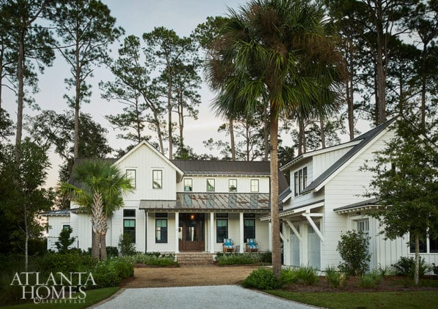 Modern farmhouse with white siding, dark metal roof, and a weathered metal porch roof. Surrounded by tall pine trees and palm trees.