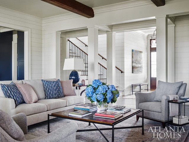 Living room with a light beige sofa and two gray armchairs, a dark wood coffee table with books and blue hydrangeas, and a view of a staircase and entryway.