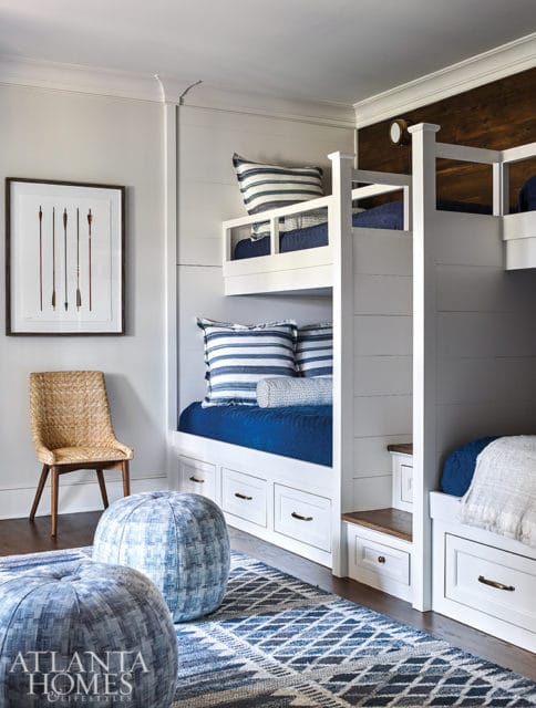 Bunk beds with white shiplap siding and navy blue bedding, accented by striped pillows and a geometric rug. A woven chair and blue poufs sit nearby.