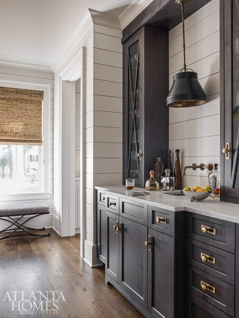 Dark wood bar cabinets with brass hardware, white marble countertop, and shiplap walls. A black pendant light hangs above the bar.