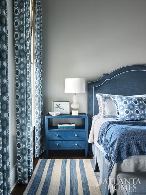 Blue upholstered headboard with white piping, bedside table with lamp and artwork, and blue patterned curtains in a bedroom.