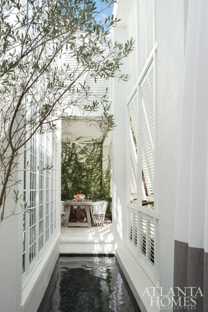 Narrow walkway with a dark water feature on the left and white shutters on the right, leading to a small patio with a wooden table, chairs, and a wall of green ivy.