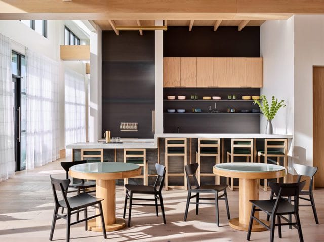 Modern kitchen with light wood cabinetry, dark gray backsplash, and white countertops. Round wooden tables with teal glass tops are surrounded by black chairs.