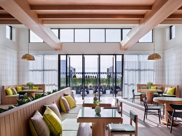 Modern lounge with light wood beams, white paneled walls, brass pendant lights, and banquette seating with lime green and patterned pillows. Glass-top tables and black chairs are arranged throughout.