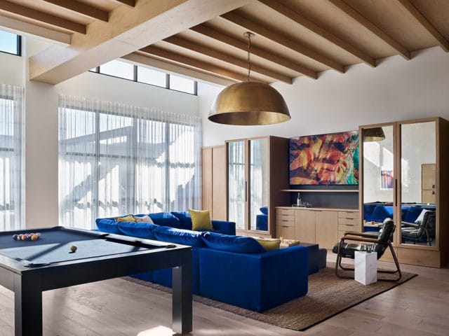 Modern living room with a royal blue sectional sofa, pool table, wood-beamed ceiling, large brass pendant light, and built-in cabinetry with mirrored doors.