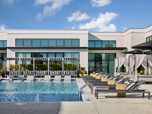 Modern building with a swimming pool and lounge chairs. White cabanas with white curtains and striped cushions are visible.