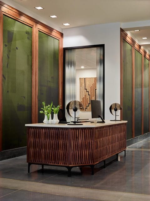 Reception desk with carved wood front and marble top, featuring two glass globe lamps, a computer monitor, and white vases with green plants.