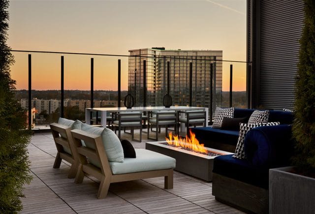 Rooftop patio with modern seating, dining table, and gas fire pit overlooking city skyline at sunset.