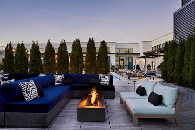 Rooftop patio with a large blue sectional sofa, modern fire pit, light blue lounge chairs, and a swimming pool with cabanas in the background.