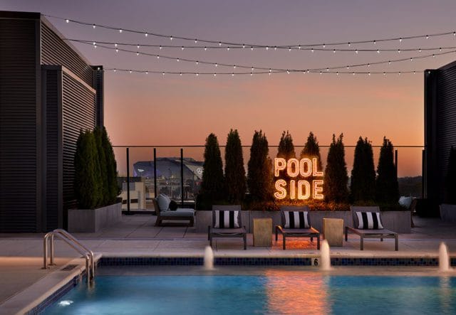 Rooftop pool area at dusk with string lights overhead, lounge chairs, and a neon 'POOL SIDE' sign. City skyline visible in the distance.