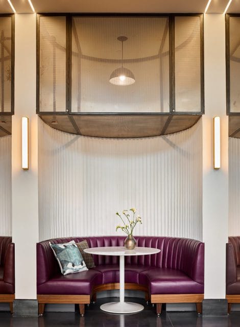 Plush purple leather banquette seating with a round white table and a vase of flowers, set against a fluted white wall and framed by mesh panels with pendant lights.