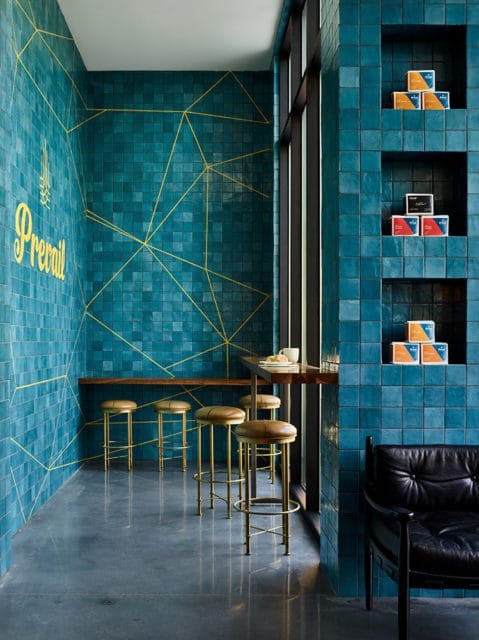 Modern cafe interior with teal square tile walls featuring gold geometric accents, a wooden counter with bar stools, and built-in shelving displaying boxes.