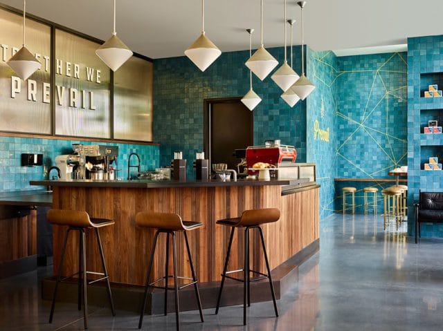 Modern coffee bar with teal tiled walls, a wooden counter, and brown leather bar stools. Text reading "TOGETHER WE PREVAIL" is visible on the wall.
