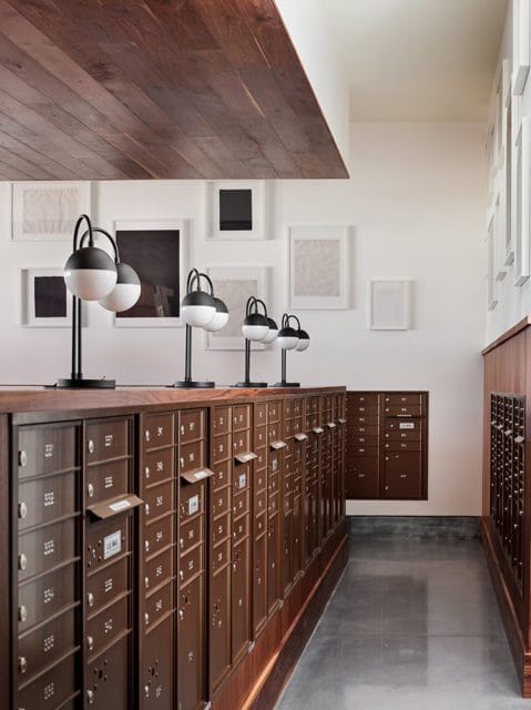 Hallway with rows of dark wood mailboxes, modern globe lamps on top, and framed art on the white wall. A slatted wood ceiling is visible.