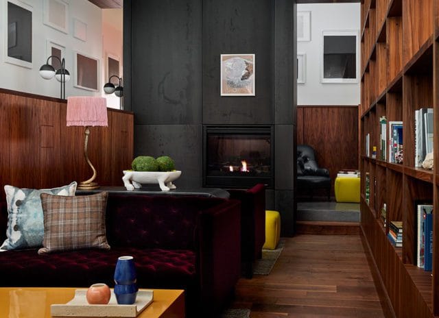Living room with a deep red tufted sofa, plaid and patterned pillows, a brass lamp with a pink fringe shade, and a fireplace with a dark stone surround.