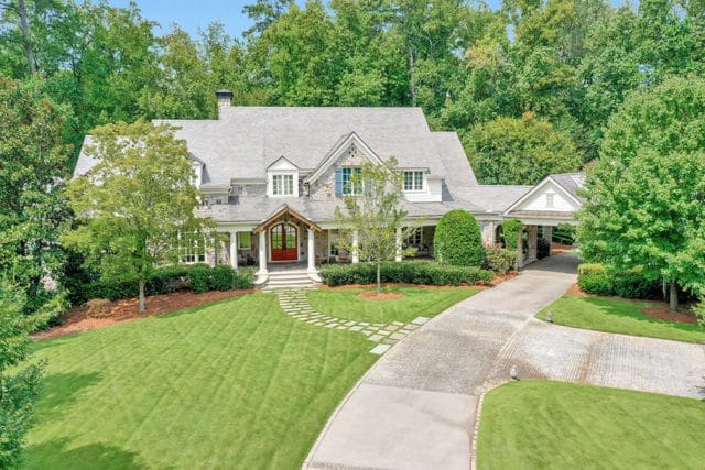Stone and shingle-sided luxury home with a red double-door entrance, covered porch, and porte-cochère, surrounded by lush green trees and manicured lawn.
