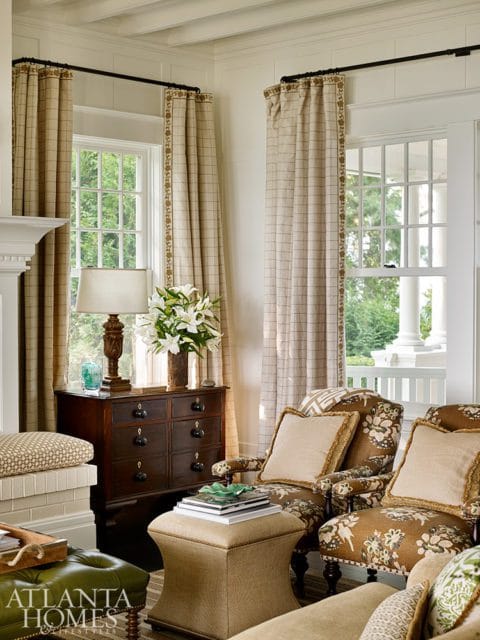 Living room with two patterned armchairs, a tufted green ottoman, and a dark wood dresser with a lamp and flowers.