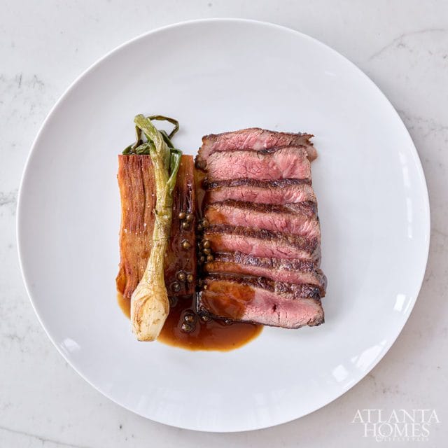 Sliced steak with sauce, roasted leeks, and peppercorns on a white plate with Atlanta Homes & Lifestyles logo in the corner