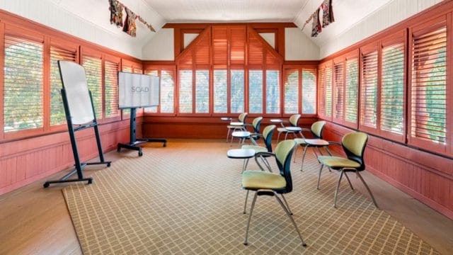 Meeting room with coral walls, wood shutters, a large screen displaying 'SCAD', a whiteboard, and modern chairs with yellow seats on a patterned carpet.