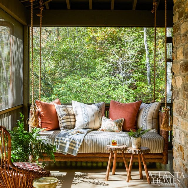 Screened porch with a wooden swing bed adorned with striped cushions, terracotta pillows, and patterned accents. A wicker chair and small log side tables complete the cozy seating area overlooking a lush forest.